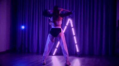 Baştan çıkarıcı dans eden seksi kadın. Titreyen bir dansçı, canlı mor ve mavi ışıkla yıkanmış, canlı neon bir üçgenin altında sahnenin enerjisini somutlaştırıyor. Bir sanatçının kadın vücudu, twerk ve yüksek topuklu ayakkabı sanatının bir kanıtı.