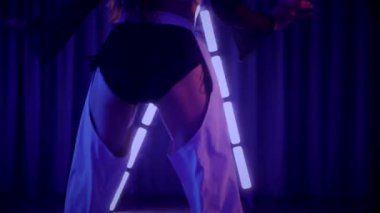 Baştan çıkarıcı dans eden seksi kadın. Titreyen bir dansçı, canlı mor ve mavi ışıkla yıkanmış, canlı neon bir üçgenin altında sahnenin enerjisini somutlaştırıyor. Bir sanatçının kadın vücudu, twerk ve yüksek topuklu ayakkabı sanatının bir kanıtı.