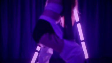 Baştan çıkarıcı dans eden seksi kadın. Titreyen bir dansçı, canlı mor ve mavi ışıkla yıkanmış, canlı neon bir üçgenin altında sahnenin enerjisini somutlaştırıyor. Bir sanatçının kadın vücudu, twerk ve yüksek topuklu ayakkabı sanatının bir kanıtı.