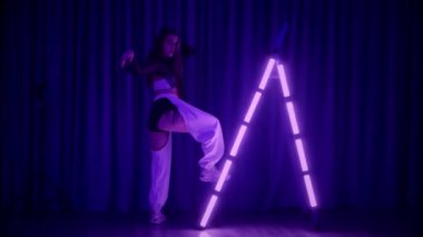 Baştan çıkarıcı dans eden seksi kadın. Titreyen bir dansçı, canlı mor ve mavi ışıkla yıkanmış, canlı neon bir üçgenin altında sahnenin enerjisini somutlaştırıyor. Bir sanatçının kadın vücudu, twerk ve yüksek topuklu ayakkabı sanatının bir kanıtı.