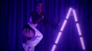 Baştan çıkarıcı dans eden seksi kadın. Titreyen bir dansçı, canlı mor ve mavi ışıkla yıkanmış, canlı neon bir üçgenin altında sahnenin enerjisini somutlaştırıyor. Bir sanatçının kadın vücudu, twerk ve yüksek topuklu ayakkabı sanatının bir kanıtı.