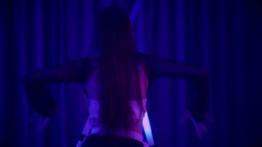 Baştan çıkarıcı dans eden seksi kadın. Titreyen bir dansçı, canlı mor ve mavi ışıkla yıkanmış, canlı neon bir üçgenin altında sahnenin enerjisini somutlaştırıyor. Bir sanatçının kadın vücudu, twerk ve yüksek topuklu ayakkabı sanatının bir kanıtı.