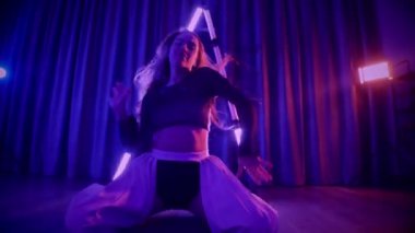 Baştan çıkarıcı dans eden seksi kadın. Titreyen bir dansçı, canlı mor ve mavi ışıkla yıkanmış, canlı neon bir üçgenin altında sahnenin enerjisini somutlaştırıyor. Bir sanatçının kadın vücudu, twerk ve yüksek topuklu ayakkabı sanatının bir kanıtı.