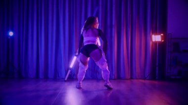 Baştan çıkarıcı dans eden seksi kadın. Titreyen bir dansçı, canlı mor ve mavi ışıkla yıkanmış, canlı neon bir üçgenin altında sahnenin enerjisini somutlaştırıyor. Bir sanatçının kadın vücudu, twerk ve yüksek topuklu ayakkabı sanatının bir kanıtı.