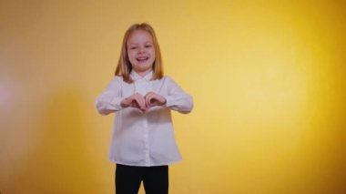 Ilık saçan genç bir kız elleriyle bir kalp oluşturur. Yüzündeki neşeli ifade, canlı bir arkaplan karşısında ışıldar. Kalp şeklinde eller, bir gülümseme ve neşeli bir mizaç. Küçük bir kızın mutluluğu, tek bir karede yakalanır.. 