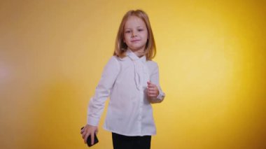 Gülümseyen kız elinde bir telefon tutuyor ve stüdyoda neşeli ve enerjik bir şekilde sarı arka planda dans ediyor. Küçük bir iş kadını stüdyoda komik bir şekilde dans ediyor..