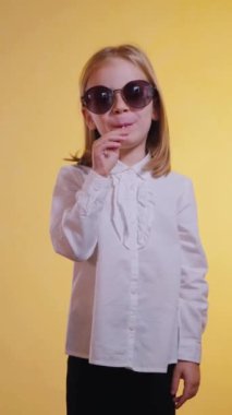 Dikey. Gıcır gıcır beyaz bir gömlek ve koyu bir pantolon giyen küçük bir kız canlı yeşil bir lolipopun tadını çıkarıyor. Yüzünde bir tatlı ve sofistike ifade var. Güneşle sırılsıklam olmuş bir arkaplanda şık bir poz