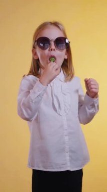 Dikey. Gıcır gıcır beyaz bir gömlek ve koyu bir pantolon giyen küçük bir kız canlı yeşil bir lolipopun tadını çıkarıyor. Yüzünde bir tatlı ve sofistike ifade var. Güneşle sırılsıklam olmuş bir arkaplanda şık bir poz