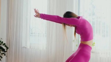 Yoga yaparken güneşli bir odada esneyen pembe elbiseli bir kadın. Gülümseyen kadın esneme egzersizleri yapar. Esneklik eğitimi, esneklik hareketleri, fiziksel egzersizler. Esneme sanatı, dinamik yoga pozu veren bir kadın. Esniyorum.