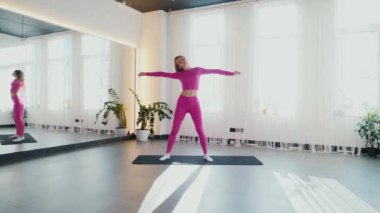 Yoga yaparken güneşli bir odada esneyen pembe elbiseli bir kadın. Gülümseyen kadın esneme egzersizleri yapar. Esneklik eğitimi, esneklik hareketleri, fiziksel egzersizler. Esneme sanatı, dinamik yoga pozu veren bir kadın. Esniyorum.