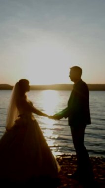 Dikey. Birbirine aşık bir çift gün batımında nehir kıyısında duruyor. Gelin ve damat el ele tutuşuyor. Öpüşen aşıkların siluetleri.