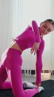 Dikey. Gülümseyen kadın esneme egzersizleri yapar. Yoga yaparken güneşli bir odada esneyen pembe elbiseli bir kadın. Esneklik eğitimi, fiziksel egzersizler.