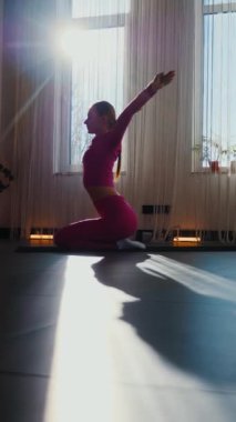 Dikey. Gülümseyen kadın esneme egzersizleri yapar. Yoga yaparken güneşli bir odada esneyen pembe elbiseli bir kadın. Esneklik eğitimi, fiziksel egzersizler.