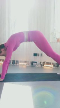Dikey. Gülümseyen kadın esneme egzersizleri yapar. Yoga yaparken güneşli bir odada esneyen pembe elbiseli bir kadın. Esneklik eğitimi, fiziksel egzersizler.