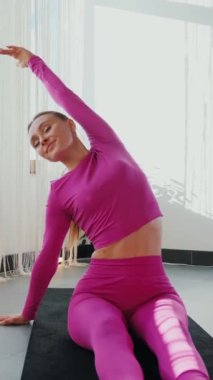 Dikey. Gülümseyen kadın esneme egzersizleri yapar. Yoga yaparken güneşli bir odada esneyen pembe elbiseli bir kadın. Esneklik eğitimi, fiziksel egzersizler.