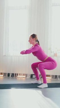 Dikey. Gülümseyen kadın esneme egzersizleri yapar. Yoga yaparken güneşli bir odada esneyen pembe elbiseli bir kadın. Esneklik eğitimi, fiziksel egzersizler.