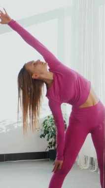 Dikey. Gülümseyen kadın esneme egzersizleri yapar. Yoga yaparken güneşli bir odada esneyen pembe elbiseli bir kadın. Esneklik eğitimi, fiziksel egzersizler.