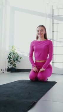 Dikey. Gülümseyen kadın esneme egzersizleri yapar. Yoga yaparken güneşli bir odada esneyen pembe elbiseli bir kadın. Esneklik eğitimi, fiziksel egzersizler.