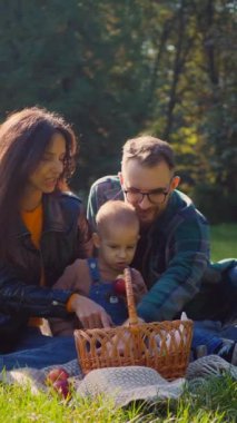 Aile pikniği, altın güneş ışığı banyosu, parkta elmaları ve kahkahaları paylaşmak. Sıcak bir sonbahar öğleden sonrasında, bir aile çimlerin üzerinde basit bir pikniğin tadını çıkarıyor. Tatlı gülümsemeler ve paylaşılan elmalar, bir ailenin neşesi.