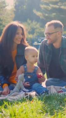 Aile pikniği, altın güneş ışığı banyosu, parkta elmaları ve kahkahaları paylaşmak. Sıcak bir sonbahar öğleden sonrasında, bir aile çimlerin üzerinde basit bir pikniğin tadını çıkarıyor. Tatlı gülümsemeler ve paylaşılan elmalar, bir ailenin neşesi.