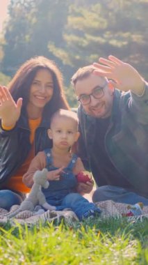 Dikey, güneşli bir öğleden sonra pikniği, kahkaha ve aşkla dolu. Aile dışarıda eğleniyor ve kameraya el sallıyor. Sıcak güneş ışığı, mutlu yüzler ve dost dalgalar. Mükemmel bir aile anı..