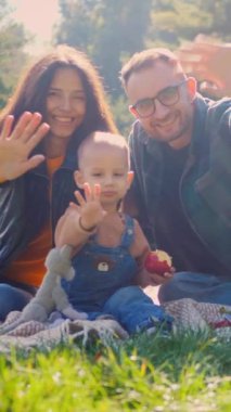 Dikey, güneşli bir öğleden sonra pikniği, kahkaha ve aşkla dolu. Aile dışarıda eğleniyor ve kameraya el sallıyor. Sıcak güneş ışığı, mutlu yüzler ve dost dalgalar. Mükemmel bir aile anı..