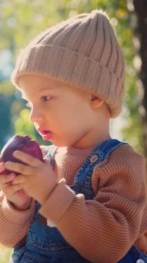 Dikey, küçük bir tanesi, ısırık bir elma tutuyor, sonbaharın altın parıltısının ortasında. Güneş ışığı, yarısı yenmiş bir elmayı tutarken çocuğun yüzünü ısıtır. Kot pantolon ve şeftali rengi bir çocuk.