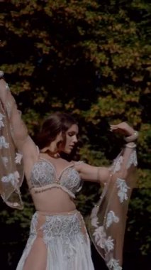 Dikey. Ormanda göbek dansı yapan bir kız. Doğulu dansı. Beyaz göbek dansı kostümlü kadın sahada hareketlerle poz veriyor. Beyaz kostümlü zarif bir dansçı. Göbek dansı yapan bir kadın. Ormanda bir baykuş dans ediyor.