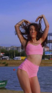 Dikey, pembe atletik şortlar ve uyumlu bir üst, bir kadın güneşin sıcaklığını kucaklıyor, poz veriyor ve güneşleniyor, saçları özgürce akıyor. Canlı pembe mayo giymiş bir kadın, kollarını açmış, deniz kenarında bir yaz gününün altın ışığında güneşleniyor.. 