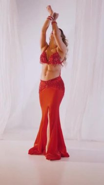 Dikey. Güzel kız oryantal göbek dansı yapıyor. Kırmızı iç çamaşırlı seksi kadın beyaz bir stüdyoda baştan çıkarıcı bir dans yapıyor. Yavaş çekim. Güzel karın ve göğüsler..