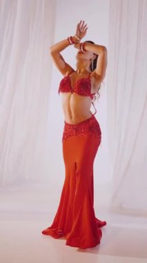 Dikey. Güzel kız oryantal göbek dansı yapıyor. Kırmızı iç çamaşırlı seksi kadın beyaz bir stüdyoda baştan çıkarıcı bir dans yapıyor. Yavaş çekim. Güzel karın ve göğüsler..