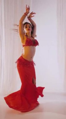 Dikey. Güzel kız oryantal göbek dansı yapıyor. Kırmızı iç çamaşırlı seksi kadın beyaz bir stüdyoda baştan çıkarıcı bir dans yapıyor. Yavaş çekim. Güzel karın ve göğüsler..