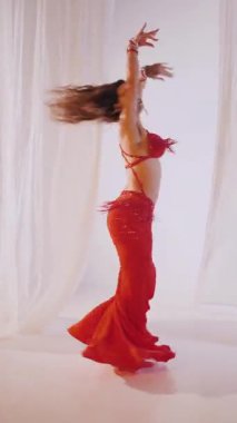 Dikey. Güzel kız oryantal göbek dansı yapıyor. Kırmızı iç çamaşırlı seksi kadın beyaz bir stüdyoda baştan çıkarıcı bir dans yapıyor. Yavaş çekim. Güzel karın ve göğüsler..