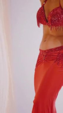 Dikey. Güzel kız oryantal göbek dansı yapıyor. Kırmızı iç çamaşırlı seksi kadın beyaz bir stüdyoda baştan çıkarıcı bir dans yapıyor. Yavaş çekim. Güzel karın ve göğüsler..
