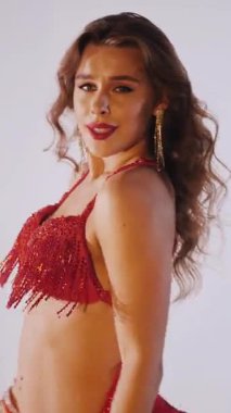 Dikey. Güzel kız oryantal göbek dansı yapıyor. Kırmızı iç çamaşırlı seksi kadın beyaz bir stüdyoda baştan çıkarıcı bir dans yapıyor. Yavaş çekim. Güzel karın ve göğüsler..