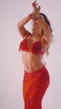 Dikey. Güzel kız oryantal göbek dansı yapıyor. Kırmızı iç çamaşırlı seksi kadın beyaz bir stüdyoda baştan çıkarıcı bir dans yapıyor. Yavaş çekim. Güzel karın ve göğüsler..