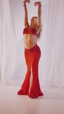 Dikey. Güzel kız oryantal göbek dansı yapıyor. Kırmızı iç çamaşırlı seksi kadın beyaz bir stüdyoda baştan çıkarıcı bir dans yapıyor. Yavaş çekim. Güzel karın ve göğüsler..
