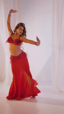 Dikey. Güzel kız oryantal göbek dansı yapıyor. Kırmızı iç çamaşırlı seksi kadın beyaz bir stüdyoda baştan çıkarıcı bir dans yapıyor. Yavaş çekim. Güzel karın ve göğüsler..