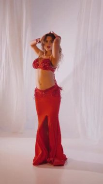 Dikey. Güzel kız oryantal göbek dansı yapıyor. Kırmızı iç çamaşırlı seksi kadın beyaz bir stüdyoda baştan çıkarıcı bir dans yapıyor. Yavaş çekim. Güzel karın ve göğüsler..