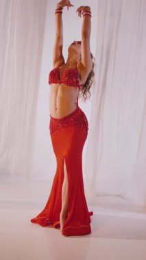 Dikey. Güzel kız oryantal göbek dansı yapıyor. Kırmızı iç çamaşırlı seksi kadın beyaz bir stüdyoda baştan çıkarıcı bir dans yapıyor. Yavaş çekim. Güzel karın ve göğüsler..