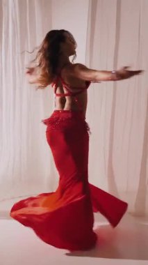 Dikey. Güzel kız oryantal göbek dansı yapıyor. Kırmızı iç çamaşırlı seksi kadın beyaz bir stüdyoda baştan çıkarıcı bir dans yapıyor. Yavaş çekim. Güzel karın ve göğüsler..