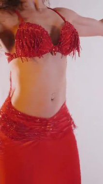 Dikey. Güzel kız oryantal göbek dansı yapıyor. Kırmızı iç çamaşırlı seksi kadın beyaz bir stüdyoda baştan çıkarıcı bir dans yapıyor. Yavaş çekim. Güzel karın ve göğüsler..