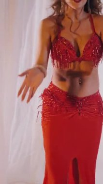 Dikey. Güzel kız oryantal göbek dansı yapıyor. Kırmızı iç çamaşırlı seksi kadın beyaz bir stüdyoda baştan çıkarıcı bir dans yapıyor. Yavaş çekim. Güzel karın ve göğüsler..