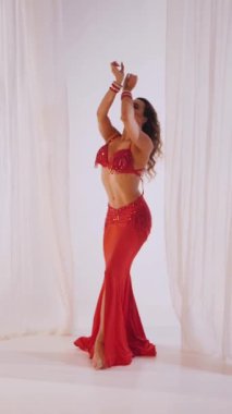 Dikey. Güzel kız oryantal göbek dansı yapıyor. Kırmızı iç çamaşırlı seksi kadın beyaz bir stüdyoda baştan çıkarıcı bir dans yapıyor. Yavaş çekim. Güzel karın ve göğüsler..