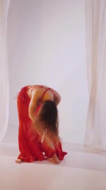 Dikey. Güzel kız oryantal göbek dansı yapıyor. Kırmızı iç çamaşırlı seksi kadın beyaz bir stüdyoda baştan çıkarıcı bir dans yapıyor. Yavaş çekim. Güzel karın ve göğüsler..