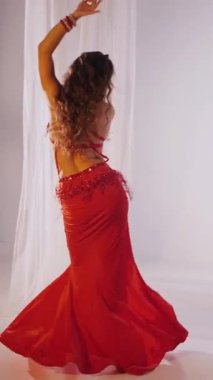 Dikey. Güzel kız oryantal göbek dansı yapıyor. Kırmızı iç çamaşırlı seksi kadın beyaz bir stüdyoda baştan çıkarıcı bir dans yapıyor. Yavaş çekim. Güzel karın ve göğüsler..