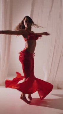 Dikey. Güzel kız oryantal göbek dansı yapıyor. Kırmızı iç çamaşırlı seksi kadın beyaz bir stüdyoda baştan çıkarıcı bir dans yapıyor. Yavaş çekim. Güzel karın ve göğüsler..