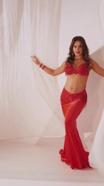 Dikey. Güzel kız oryantal göbek dansı yapıyor. Kırmızı iç çamaşırlı seksi kadın beyaz bir stüdyoda baştan çıkarıcı bir dans yapıyor. Yavaş çekim. Güzel karın ve göğüsler..