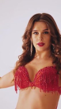 Dikey. Güzel kız oryantal göbek dansı yapıyor. Kırmızı iç çamaşırlı seksi kadın beyaz bir stüdyoda baştan çıkarıcı bir dans yapıyor. Yavaş çekim. Güzel karın ve göğüsler..