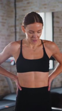 Dikey. Atletik giyinmiş genç bir kadın parlak bir spor salonunda dikiliyor. Spor salonunda kararlı görünen bir kadın. Spor salonundaki antrenmana ara veren zinde bir kadın. Gülümsüyor